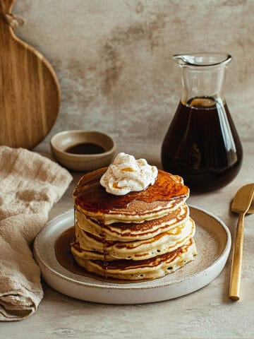 Stack of brown sugar protein pancakes topped with whipped cream and a drizzle of syrup on a ceramic plate