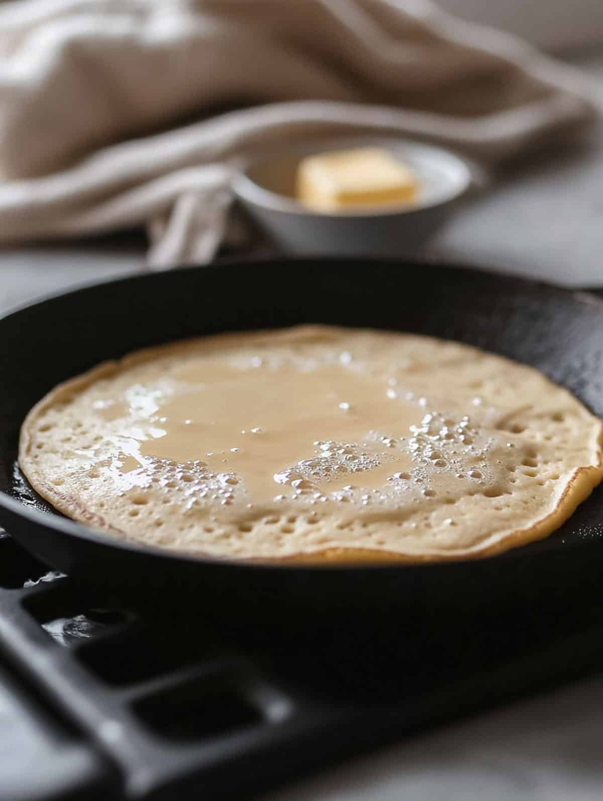 Brown sugar protein pancake with bubbles on a hot skillet, ready to be flipped, with a bowl of butter in the background.