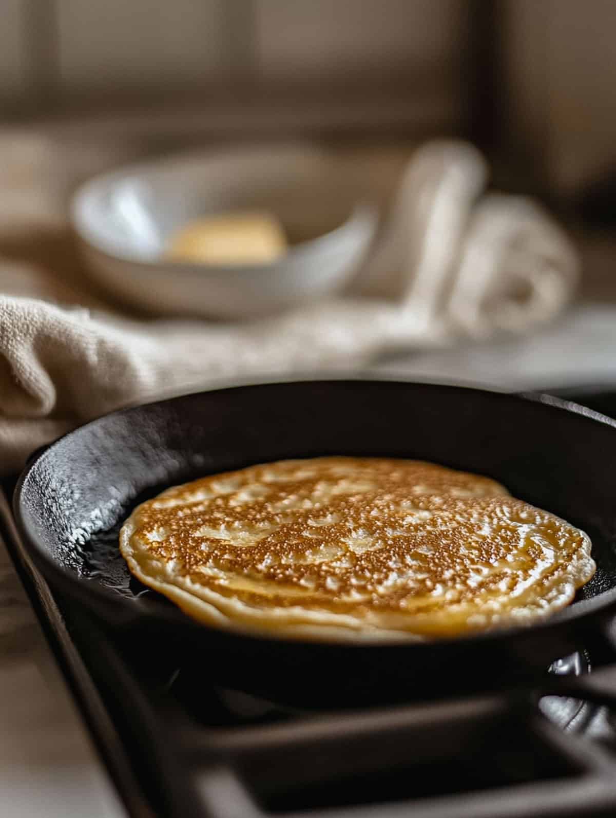 Golden-brown brown sugar protein pancake cooking in a cast iron skillet with a blurred butter dish in the background.