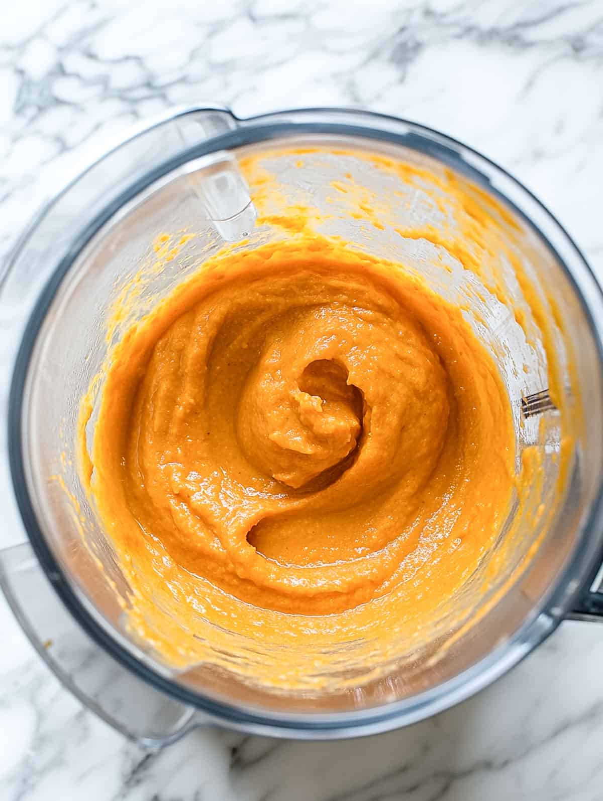 A blender on a marble counter blending cooked vegetables into a smooth, vibrant orange puree.