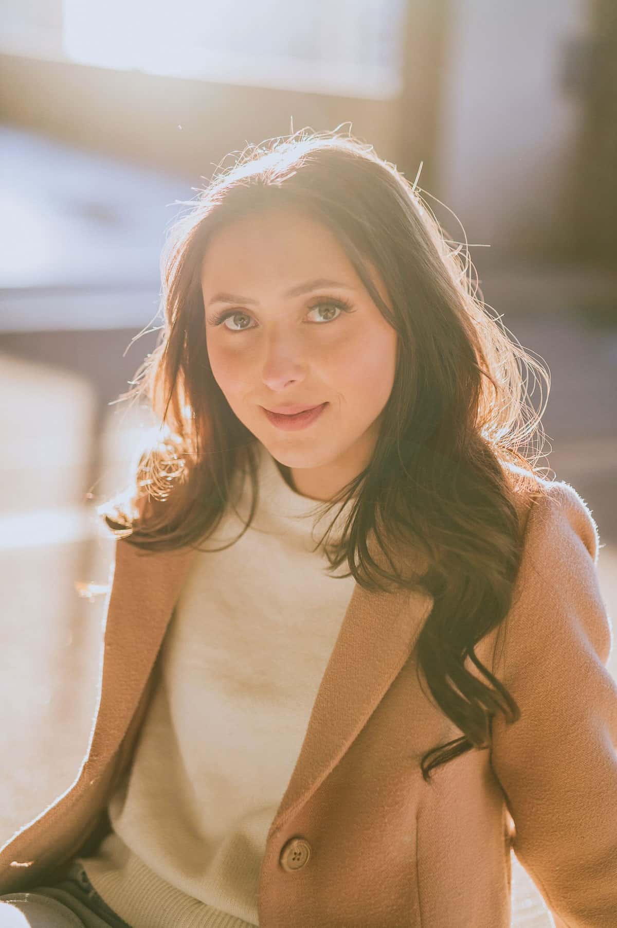 Taylor McCain, author and chef, is sitting in a warmly lit setting. She has long, wavy hair and is wearing a light-colored sweater under a tan coat. Taylor is smiling gently at the camera, with soft sunlight illuminating her face and hair, creating a serene and inviting atmosphere.