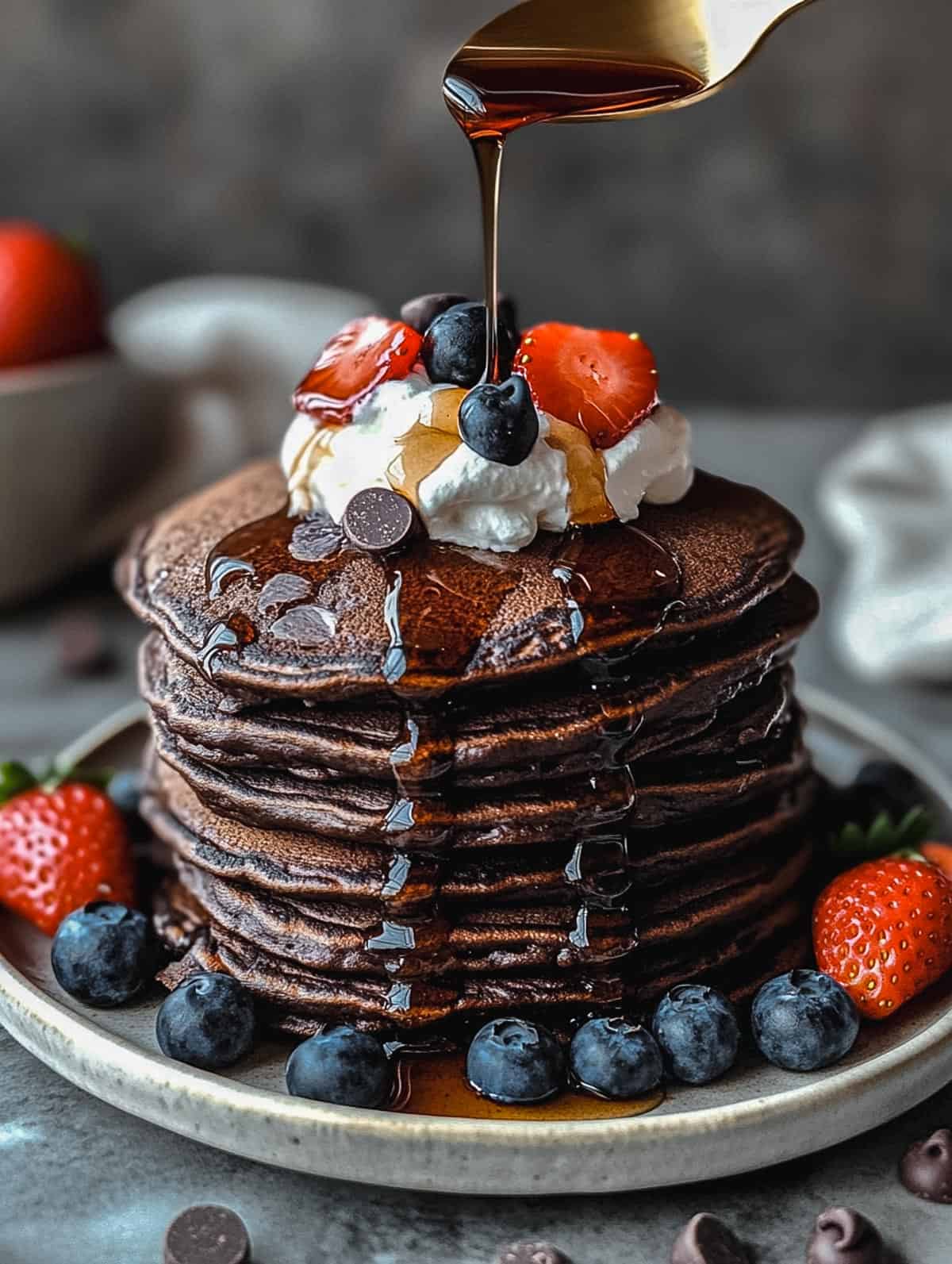 Stack of chocolate protein pancakes topped with berries, whipped cream, and syrup drizzle.