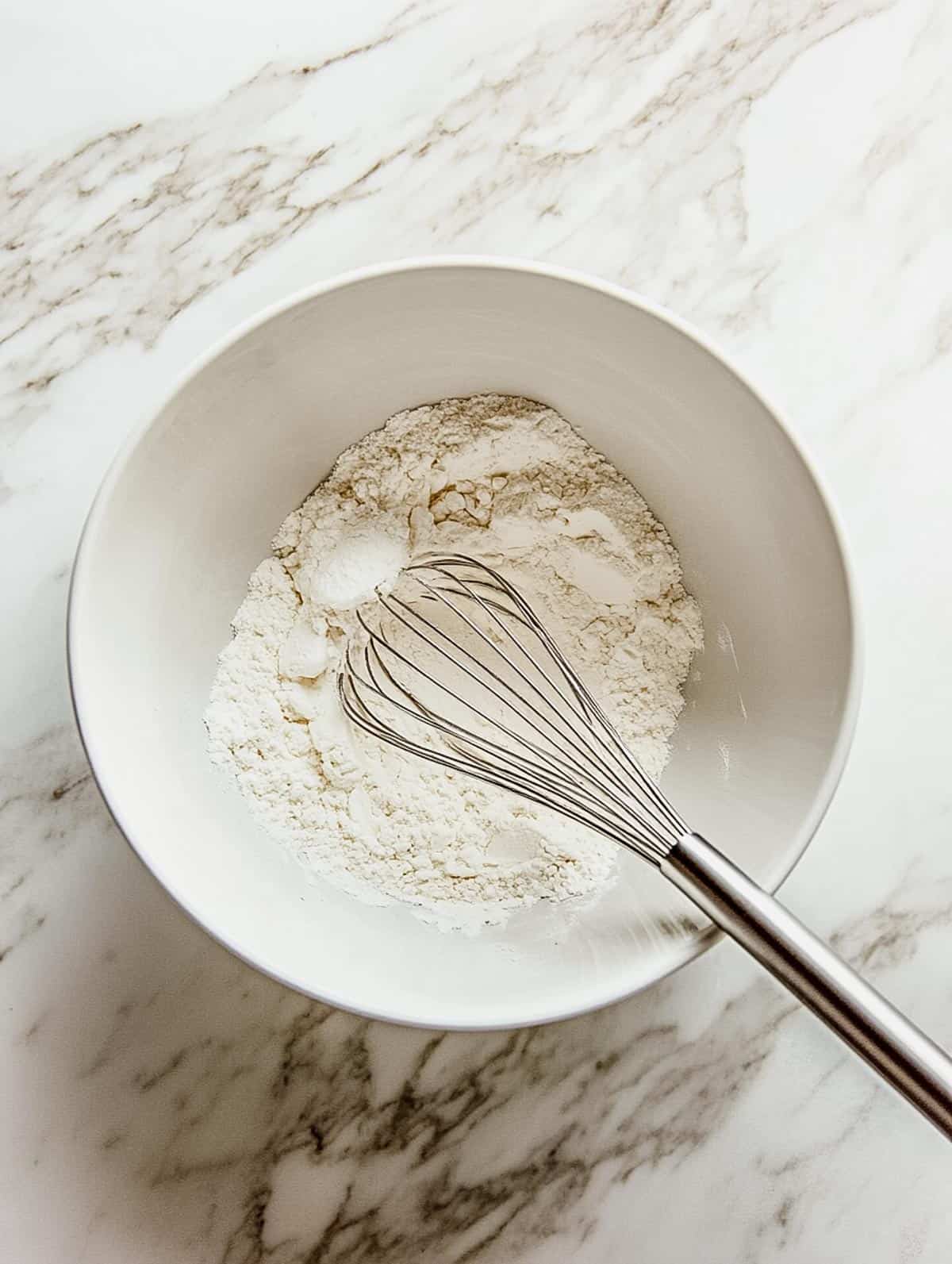 White bowl with dry ingredients and a whisk, ready to be mixed for pancake batter.