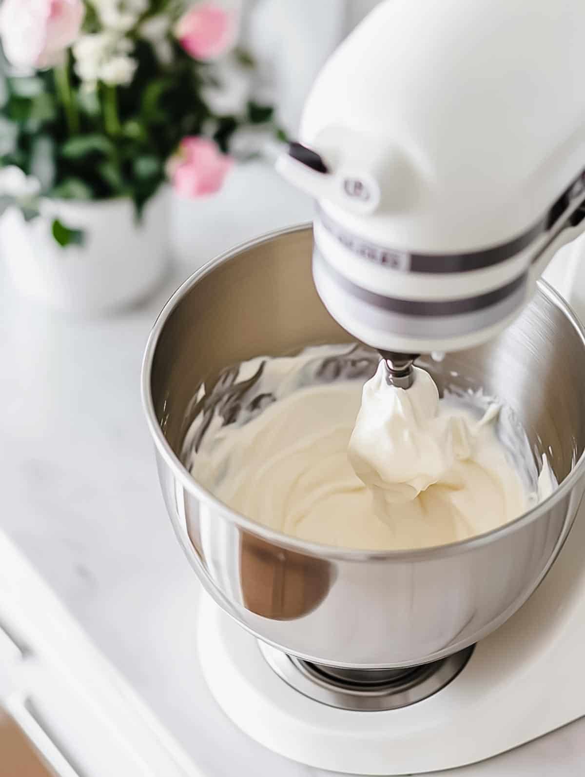 A stand mixer whisking heavy cream in a metal bowl, creating light and fluffy whipped cream, with flowers in the background.