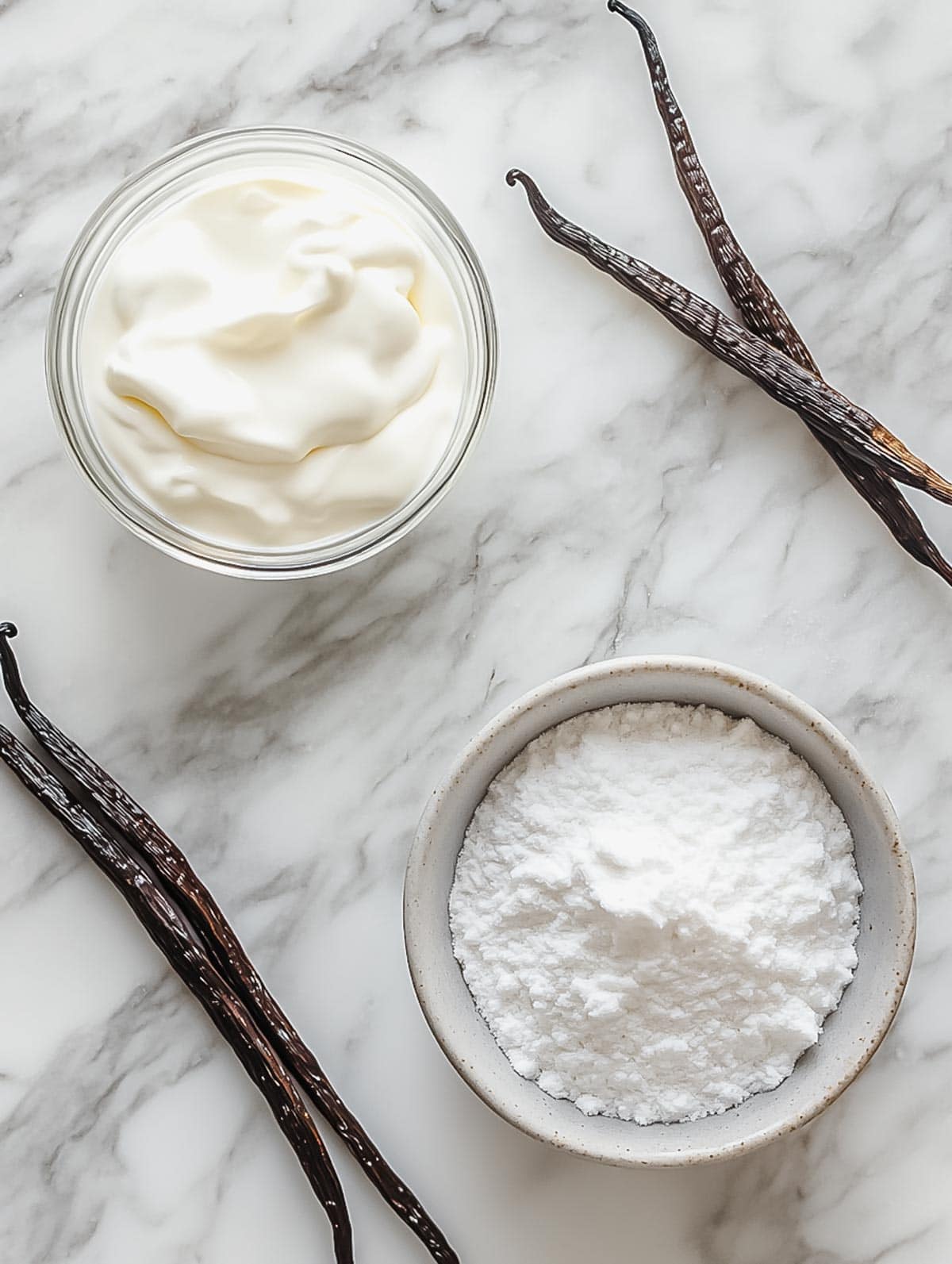 A flat lay of whipped cream ingredients, including a small bowl of powdered sugar, a bowl of cream, and vanilla beans on a marble surface.