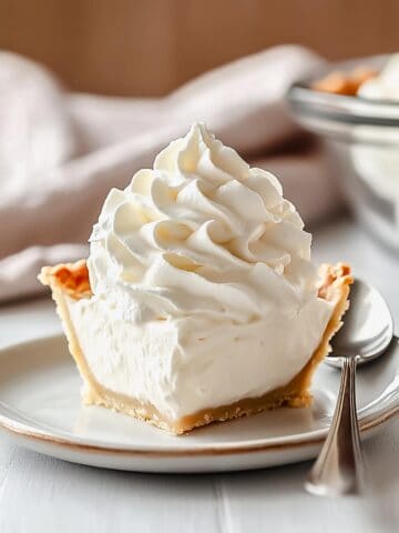 A slice of pie topped with a generous swirl of fresh whipped cream, served on a white plate with a spoon.