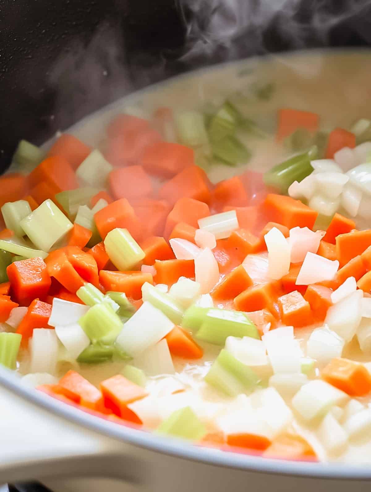 A pot filled with sautéed carrots, celery, and onions, with steam rising as the vegetables soften.