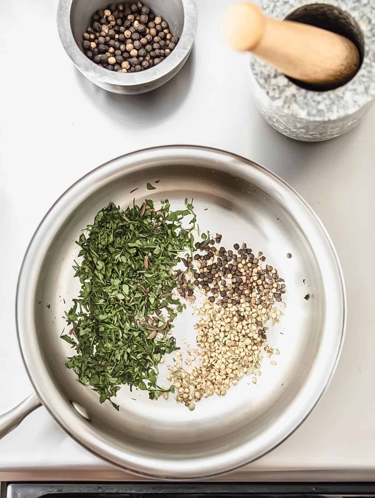 Herbs and spices being toasted in a pan, with a mortar and pestle nearby.